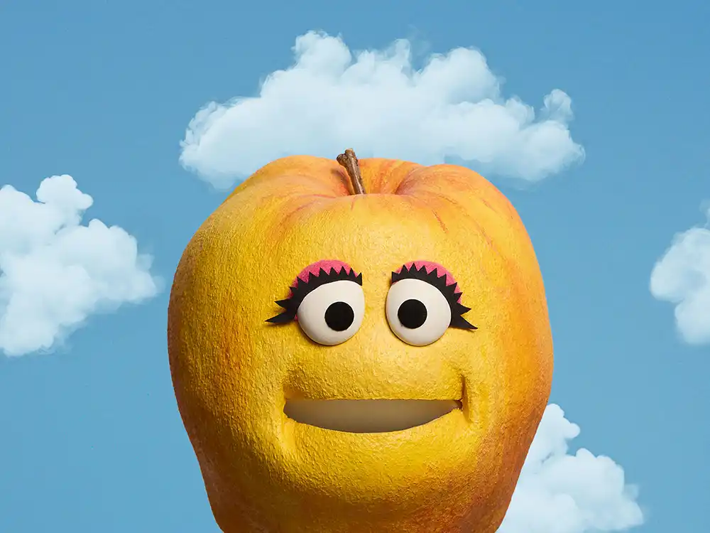 A smiling yellow apple puppet with lashes fills the frame, from Compleat x Elmo's What Is Better Than How