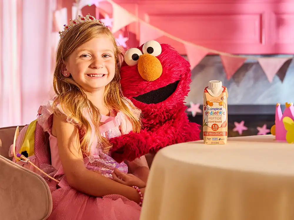 A girl in a pink princess dress sits with Elmo and Compleat Pediatric Peptide, from What Is Better Than How