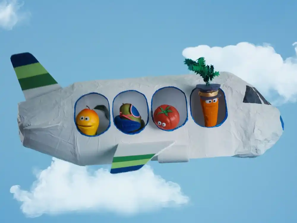 Fruit and vegetable puppets ride in a paper airplane, carrot as pilot, from Compleat x Elmo's What Is Better Than How