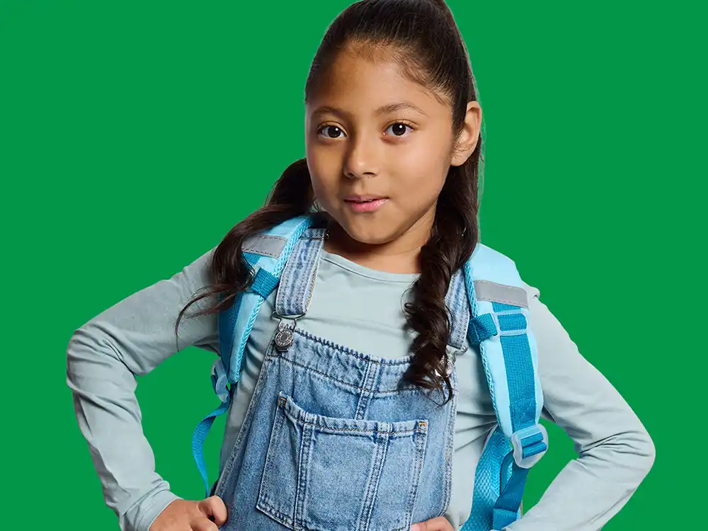 A girl in denim overalls and a blue feeding backpack stands with hands on hips, from Compleat x Elmo's What Is Better Than How