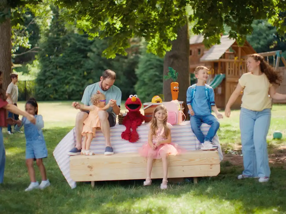Families gather with Elmo on a picnic platform under trees, from Compleat x Elmo's What Is Better Than How