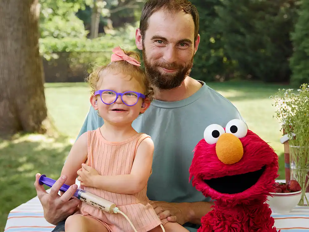 A dad holds a toddler with purple glasses and a feeding syringe beside Elmo, from Compleat x Elmo's What Is Better Than How