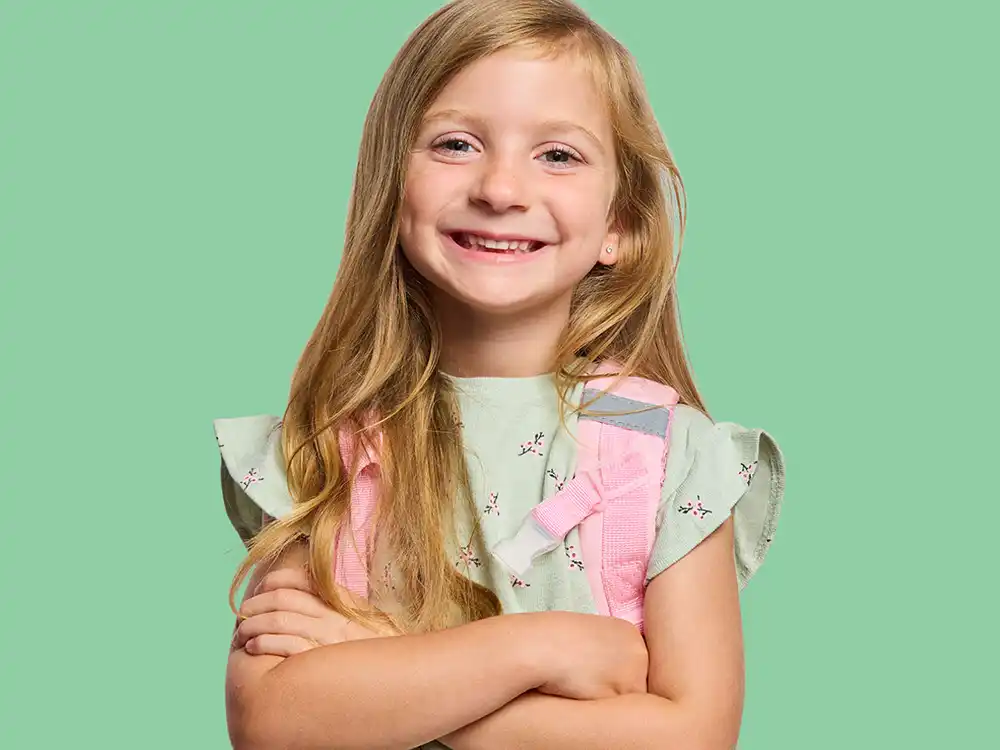 A smiling girl crosses her arms in a pink feeding backpack, from Compleat x Elmo's What Is Better Than How
