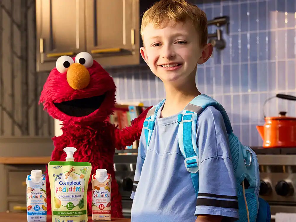 A smiling boy in a blue feeding backpack stands with Elmo and Compleat Pediatric, from What Is Better Than How