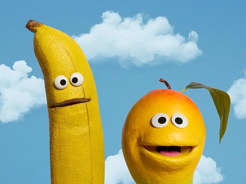 A banana puppet and a mango puppet grin against a cloudy blue sky, from Compleat x Elmo's What Is Better Than How