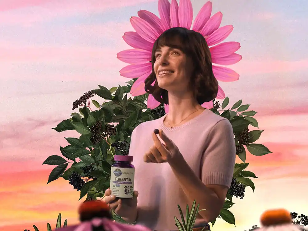A woman holds an Elderberry bottle and a gummy before a pink daisy at sunset, from Garden of Life's Formulas for Feeling Alive