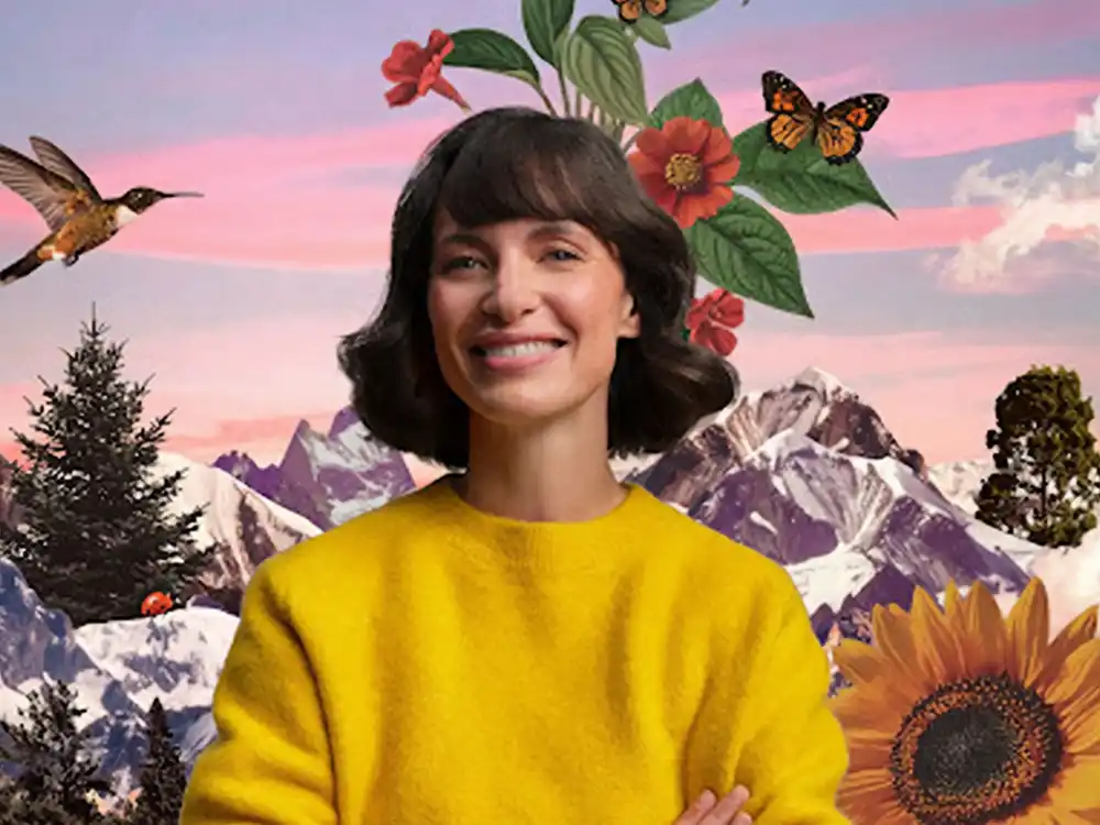 A smiling woman in a yellow sweater stands before mountains, flowers, and a hummingbird, from Garden of Life's Formulas for Feeling Alive
