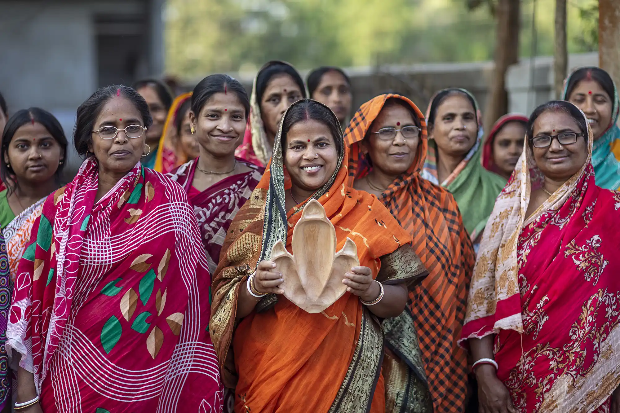 Woman in an orange sari holds a carved wooden dinosaur-footprint tray, standing among other women in colorful saris