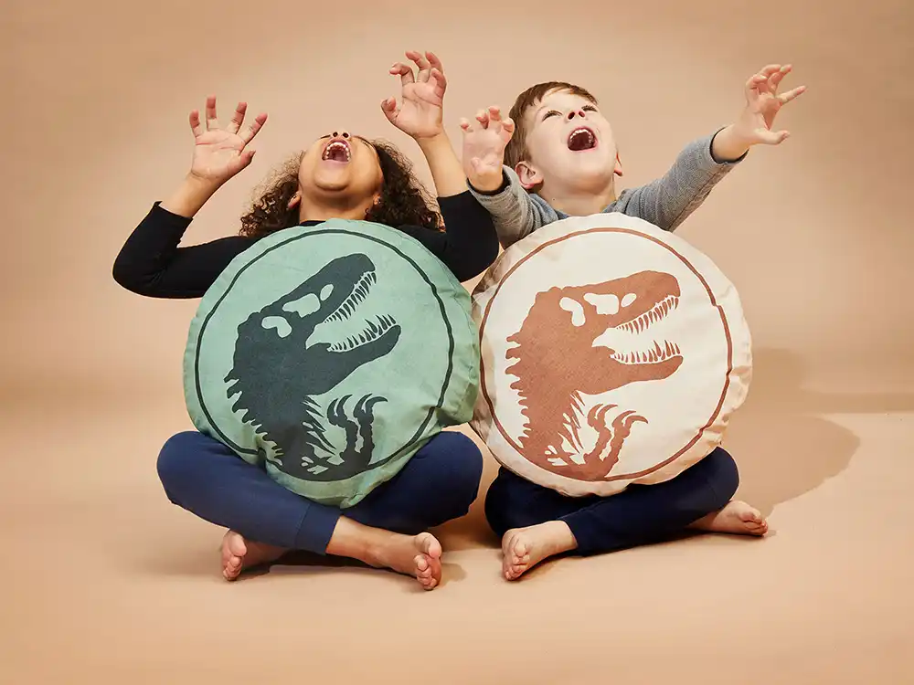 Two children sit hugging round T. rex logo pillows with arms raised, from Jurassic World's Preserving Legacies of the Past collection