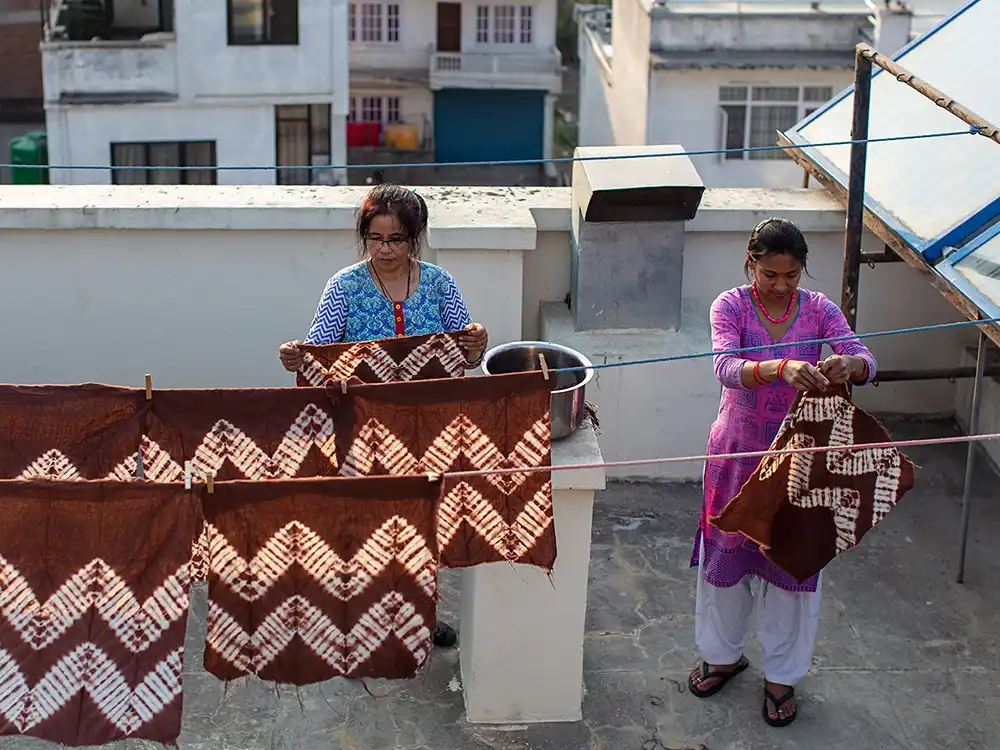 Two women hang brown-and-white patterned textiles on a rooftop clothesline, from Jurassic World's Preserving Legacies of the Past collection