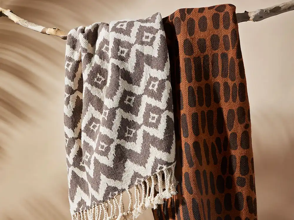 Two woven throws hang from a branch, one geometric gray-and-cream, one rust with dark ovals, from Jurassic World's Preserving Legacies of the Past collection