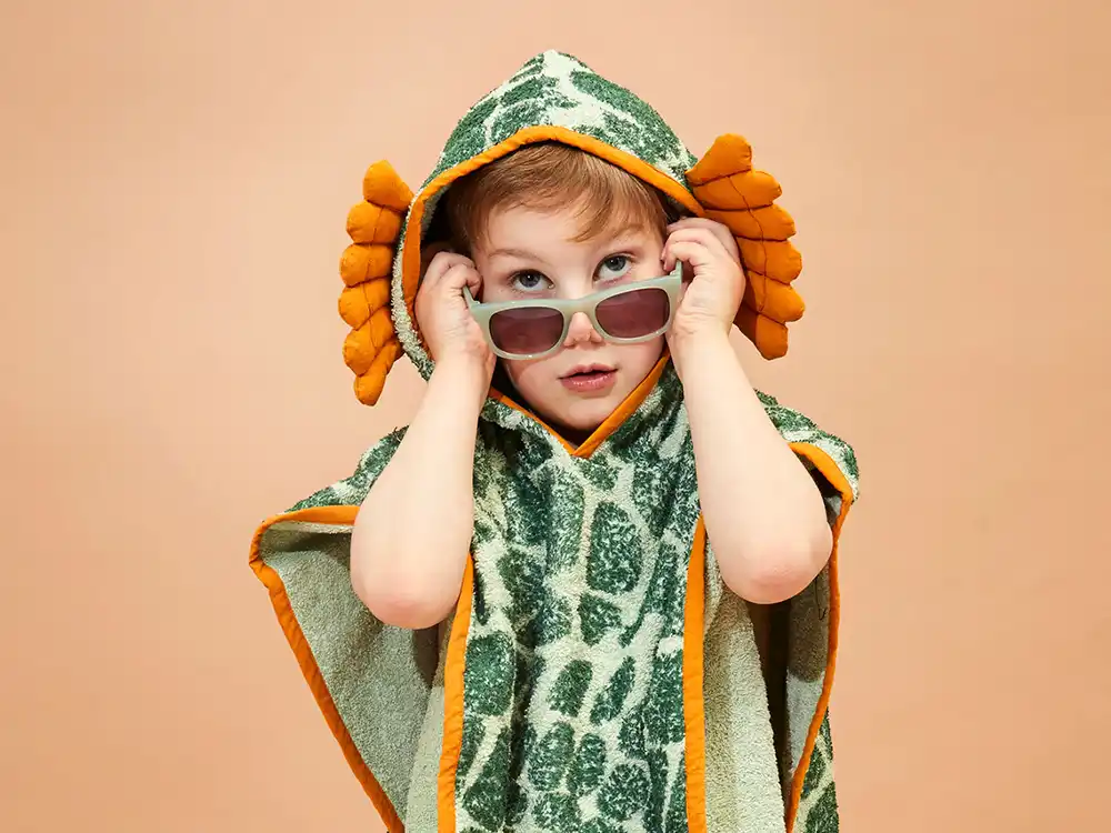Child in a green-and-orange dinosaur hooded towel holds sunglasses, from Jurassic World's Preserving Legacies of the Past collection
