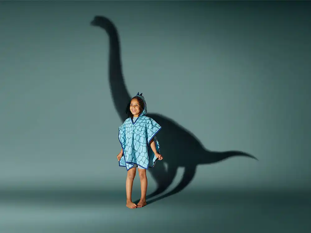 Child in a blue dinosaur hooded towel stands before a long-neck dinosaur shadow, from Jurassic World's Preserving Legacies of the Past collection