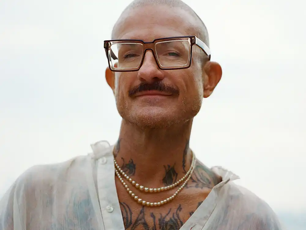 Bald man with tortoise-shell glasses, a mustache, tattoos, and pearl necklaces, from Lambda Legal's All Rise campaign
