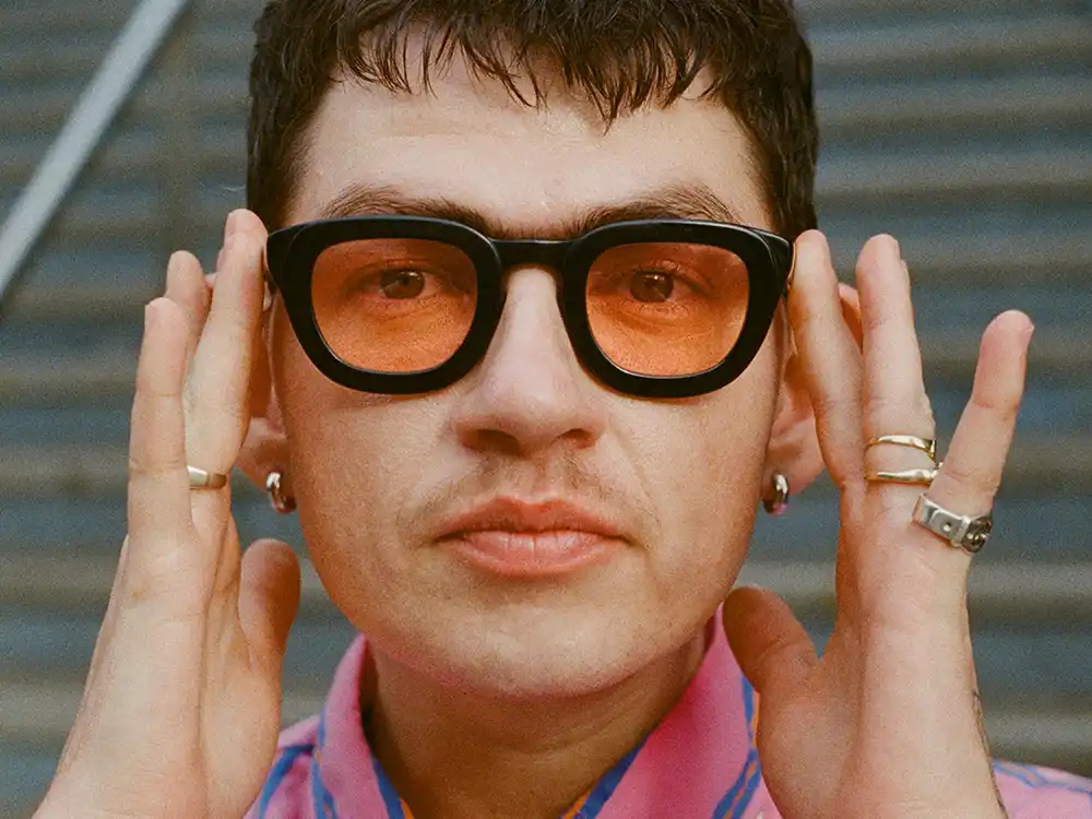 Person with a bowl cut holds thick black glasses with orange lenses, from Lambda Legal's All Rise campaign