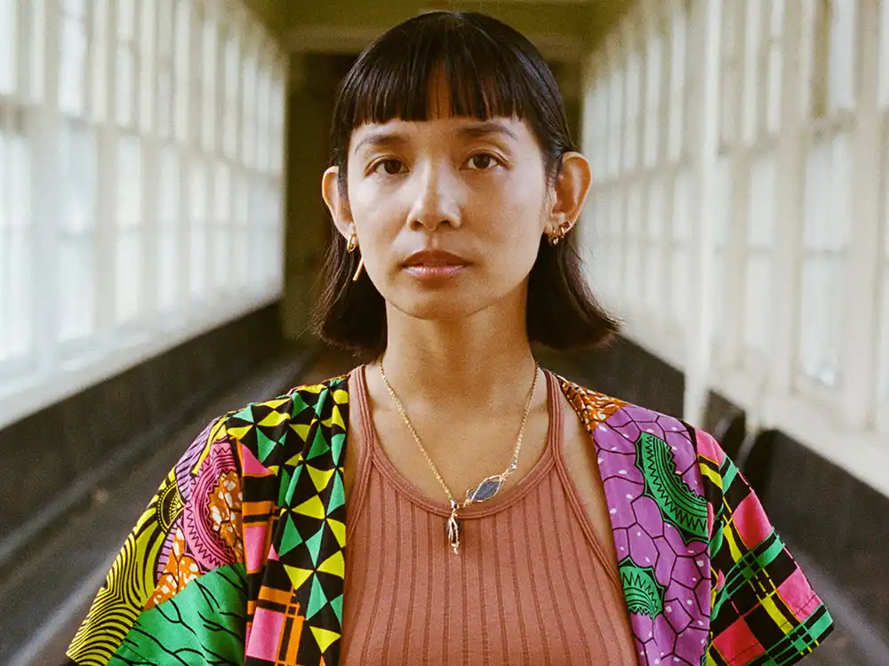 Person with a black bob and bangs wears a colorful geometric jacket in a sunlit hallway, from Lambda Legal's All Rise campaign