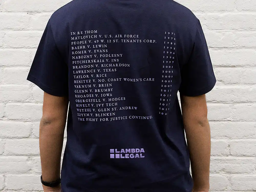 Navy T-shirt back lists LGBTQ+ case names and years above a Lambda Legal logo, from Lambda Legal's All Rise campaign