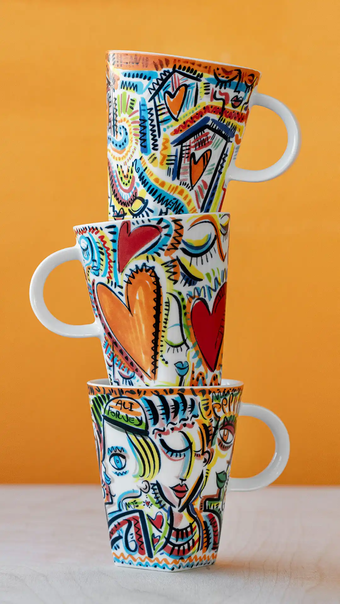 Three stacked mugs painted with colorful faces and hearts against an orange background