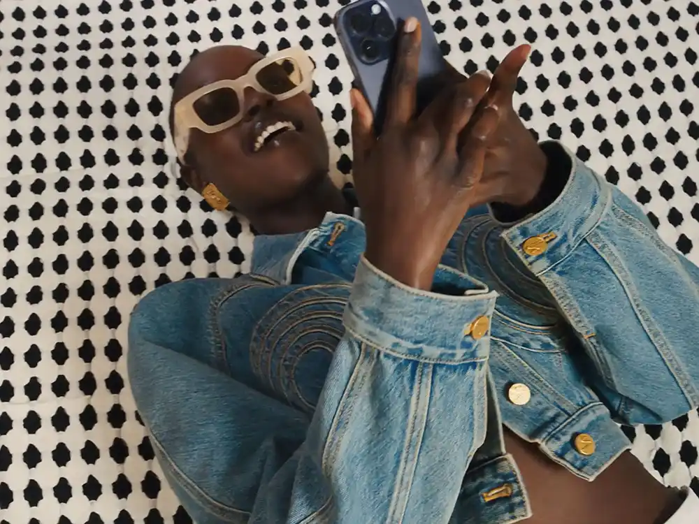 Person in cream sunglasses and a denim jacket lies on a black-and-white pattern scrolling a phone, from Vestiaire Collective's Parlez-vous?