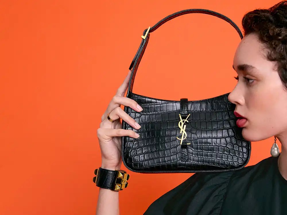 Woman holds a black YSL crocodile-texture shoulder bag beside her face, from Vestiaire Collective's Parlez-vous?
