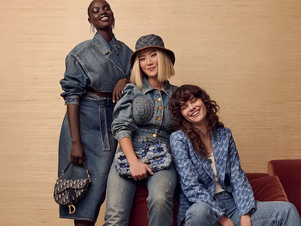 Three smiling women in denim looks pose together, one holding a Dior saddle bag, from Vestiaire Collective's Parlez-vous?