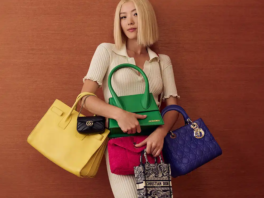Woman in a cream knit holds a stack of luxury handbags including Jacquemus and Dior, from Vestiaire Collective's Parlez-vous?