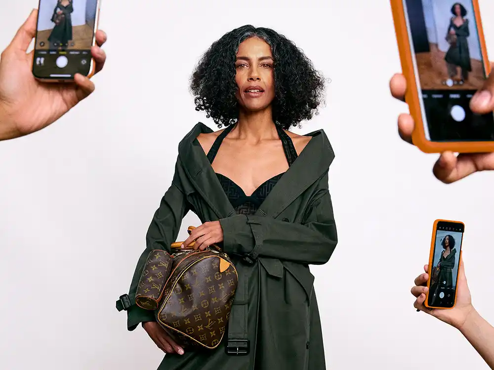 Woman in a green trench holds a Louis Vuitton Speedy as phones photograph her, from Vestiaire Collective's Parlez-vous?