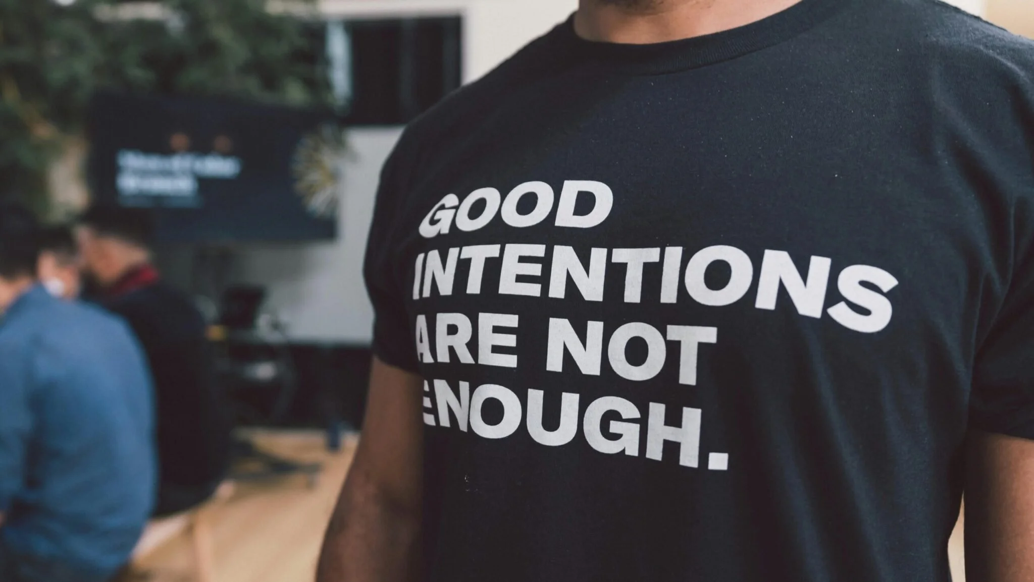 A black T-shirt reading GOOD INTENTIONS ARE NOT ENOUGH, worn in an office with colleagues in the background