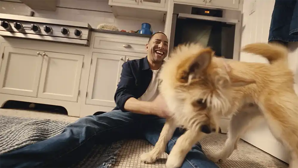 A man laughs on a kitchen floor as a fluffy dog jumps over his legs, from Native Pet's Let Dogs Be Dogs in The Drum